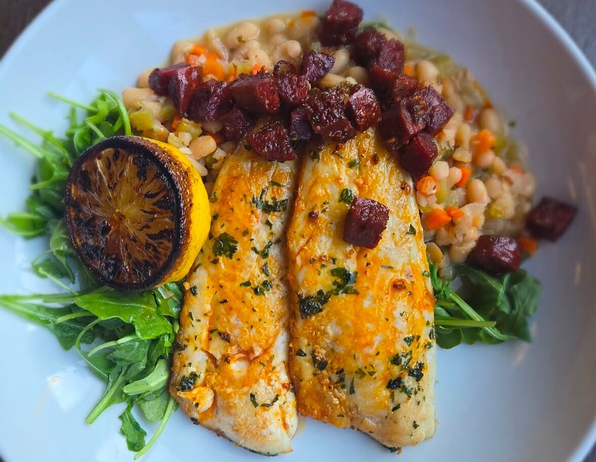 A plated gourmet fish dish featuring two golden-seared salmon fillets topped with fresh herbs, roasted beets, and chickpeas served on a bed of microgreens with a charred lemon slice, presented on a white plate.