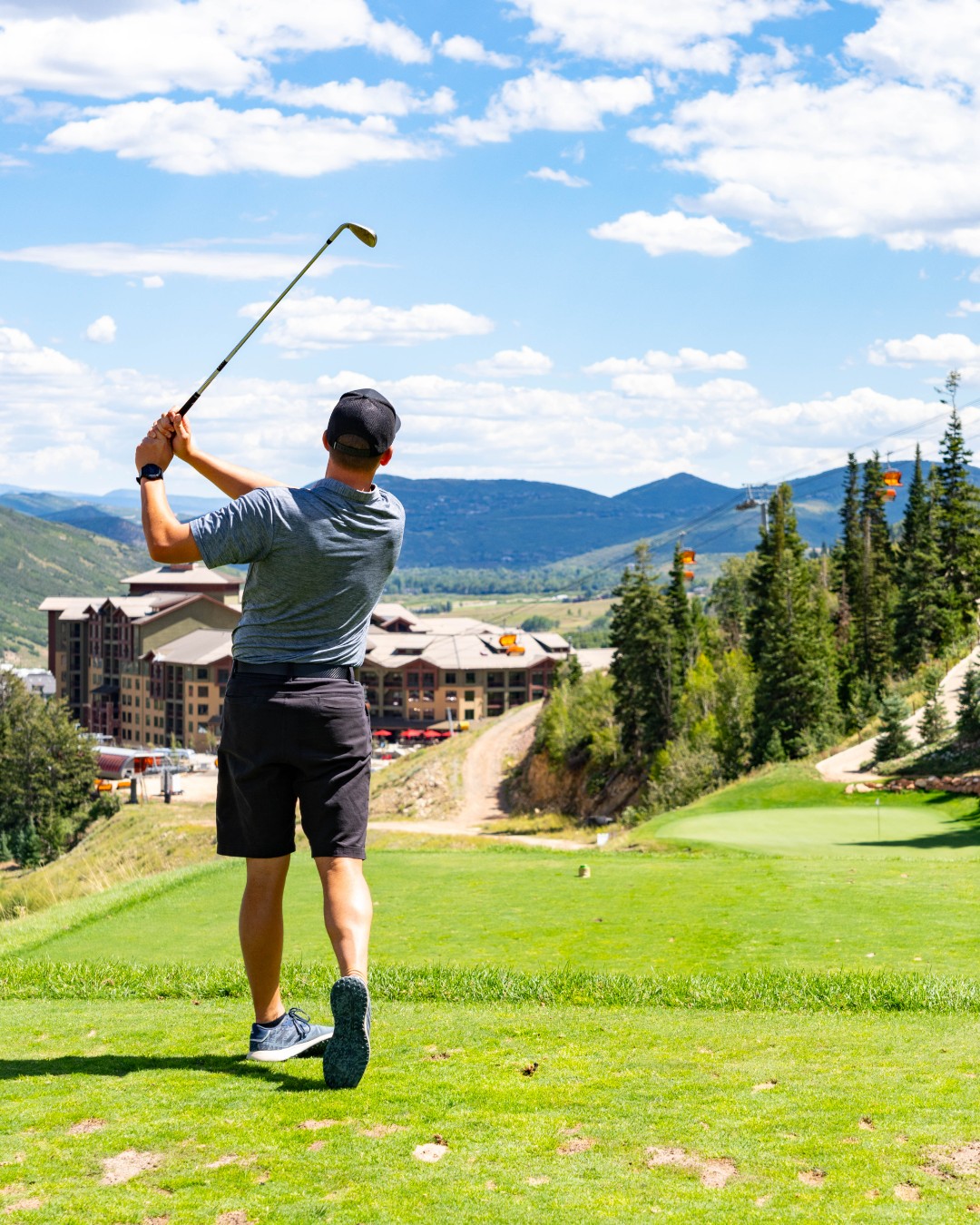 A golfer swings toward a distant green with mountains, trees, and resort buildings spread out under a bright, partly cloudy sky.