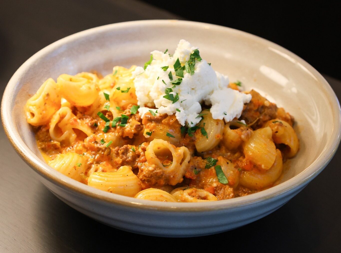 A creamy curried pasta dish featuring cavatappi noodles in a rich orange sauce topped with tender chunks of meat, garnished with fresh green herbs and a dollop of white sour cream, served in a shallow white ceramic bowl.