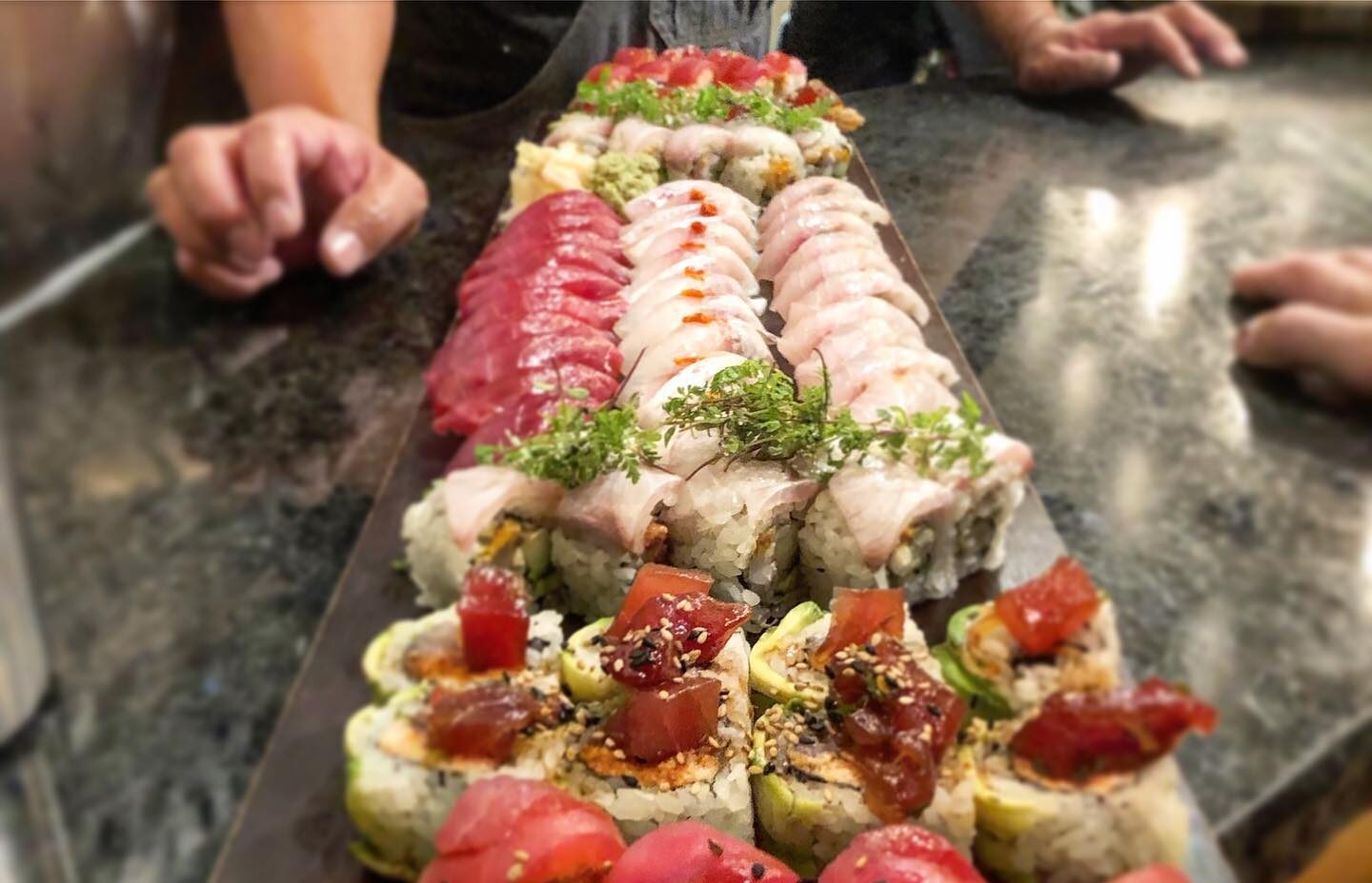 An assorted sushi roll platter displayed on a dark slate board featuring multiple varieties including red tuna rolls, white fish rolls, and spicy yellowish rolls topped with sesame seeds.